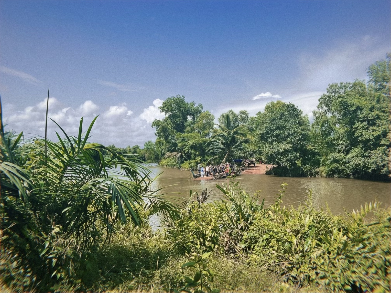 A river crossing in Cambodia, with people boarding a small ferry under palm trees.