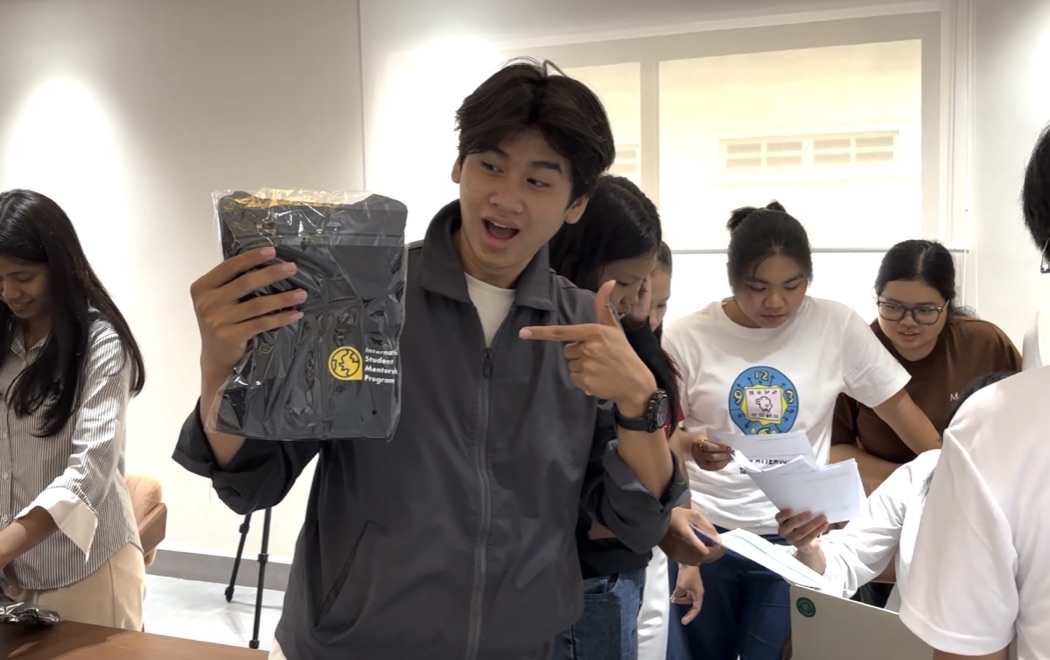 An ambassador grinning and pointing at his ISMP Ambassador jacket, still sealed in its packaging, while the rest of the cohort opens theirs.