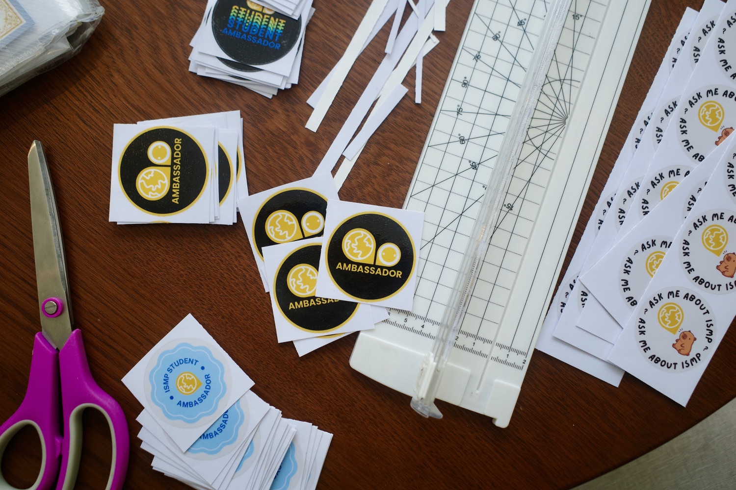 ISMP stickers mid-cut on a table — Ambassador badges and Ask me about ISMP capybara stickers beside scissors and a paper trimmer.