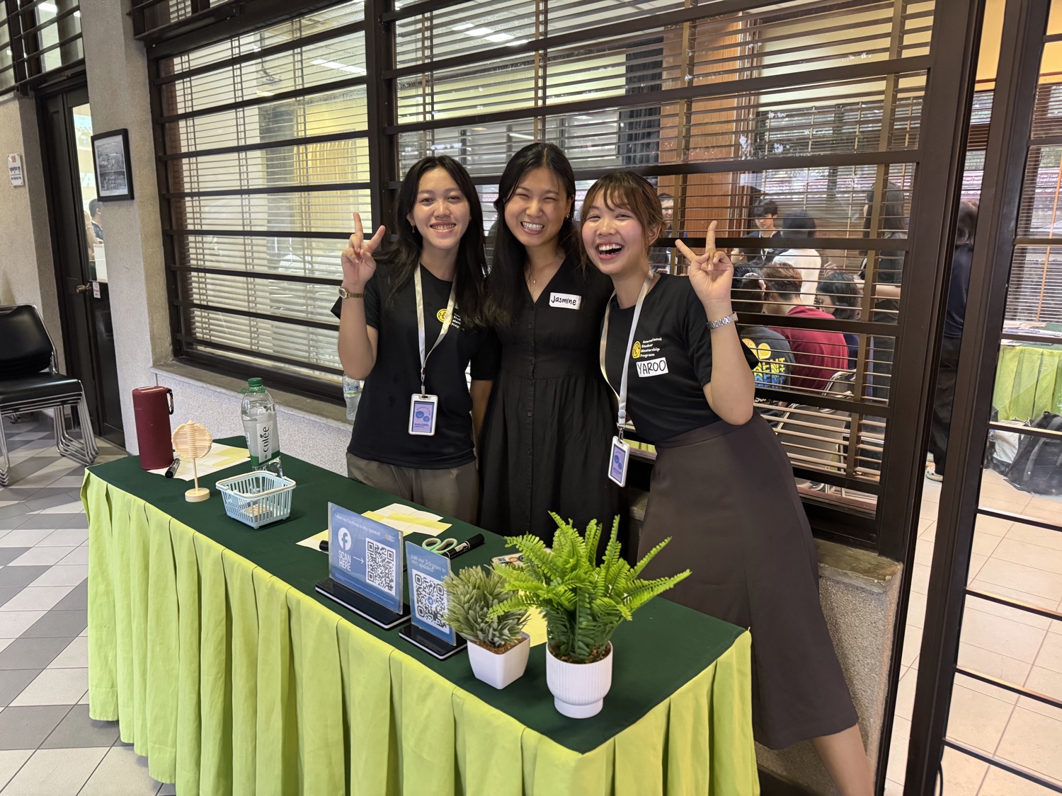 Jasmine with two ambassadors behind the welcome table with QR-code sign-up signs and plants.