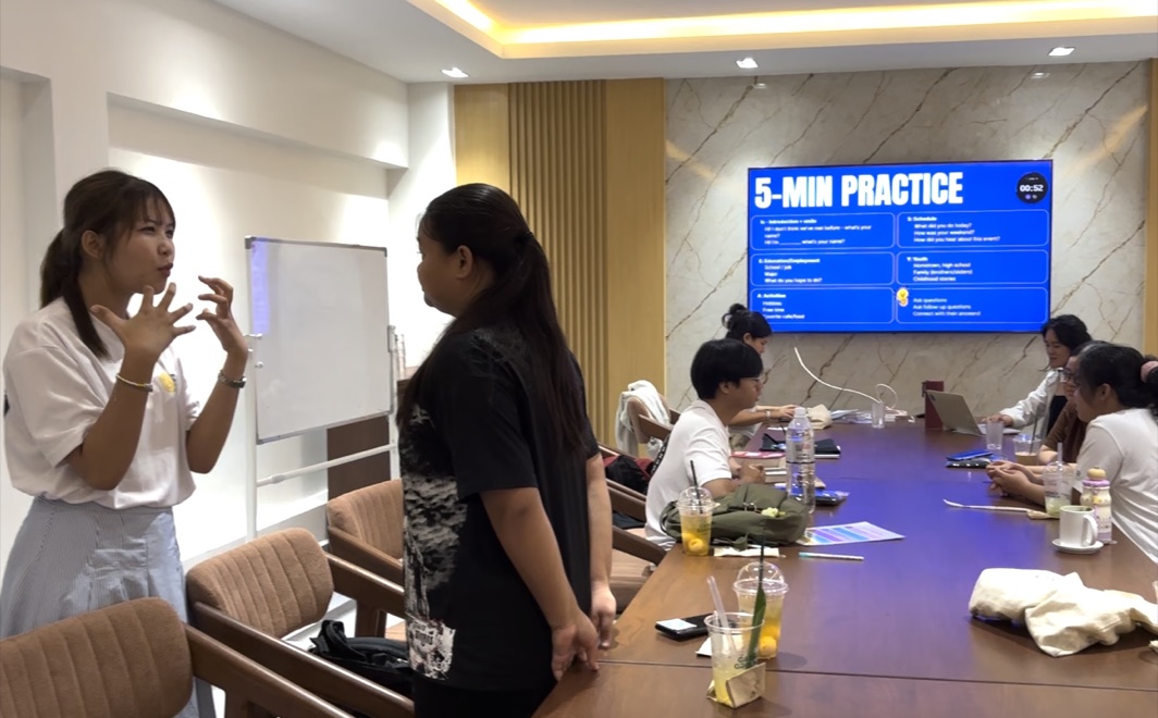 A mentor coaching a student through a five-minute conversation practice, with the prompt slide on the screen behind them.