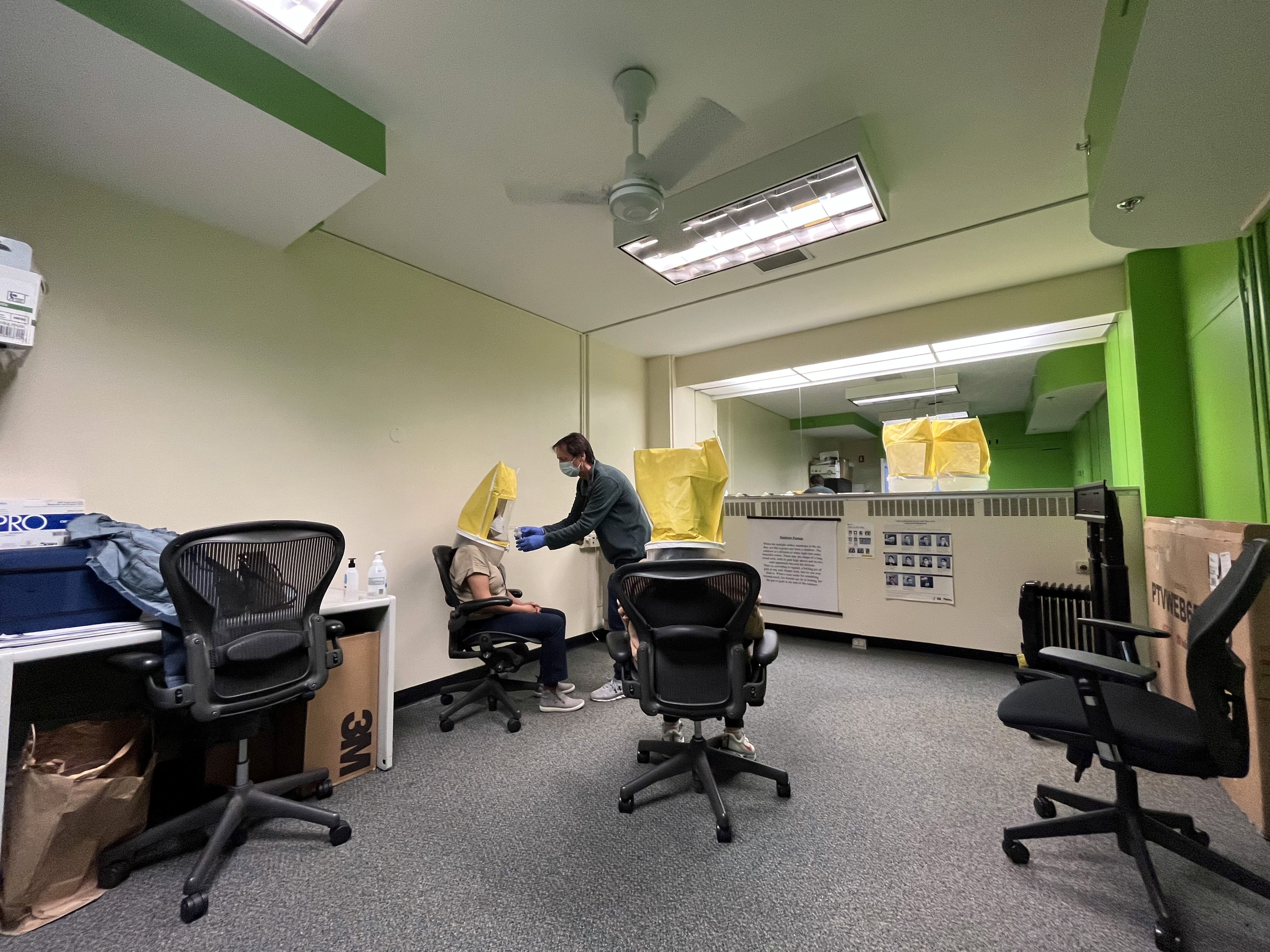 A respirator fit test session in progress: a participant wearing a yellow test hood while a masked tester administers the test, with mirrors, equipment boxes, and office chairs around the room.